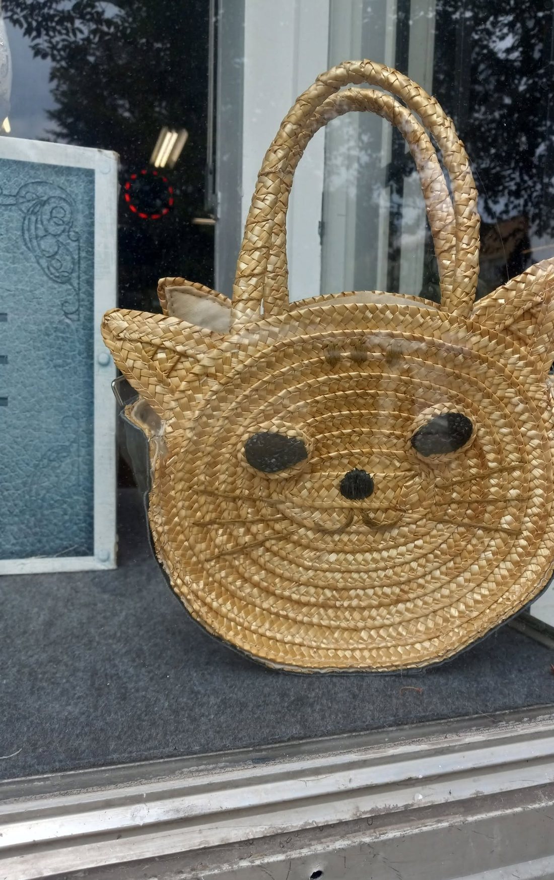A woven straw handbag shaped like a cat's head, with dark stitched eyes and a nose, propped in a shop window.