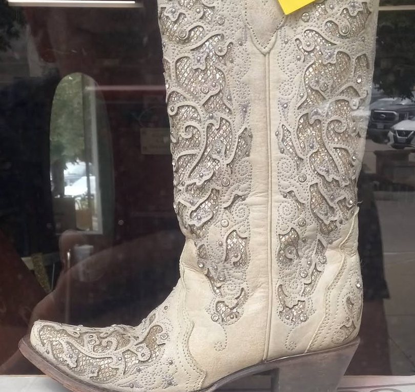Close view of a cream western boot with cut-out scrollwork and a stacked leather heel, standing behind glass.