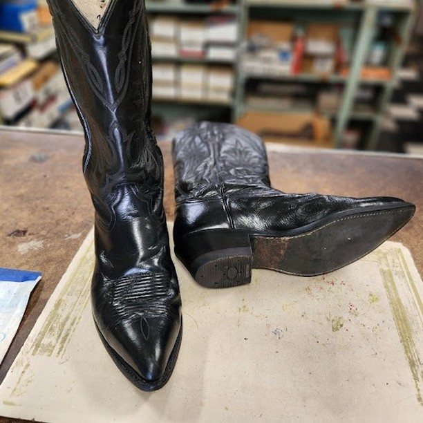 A pair of polished black cowboy boots standing on the work counter at Mace Shoe Repair, shelves of boxed and ticketed work behind them.