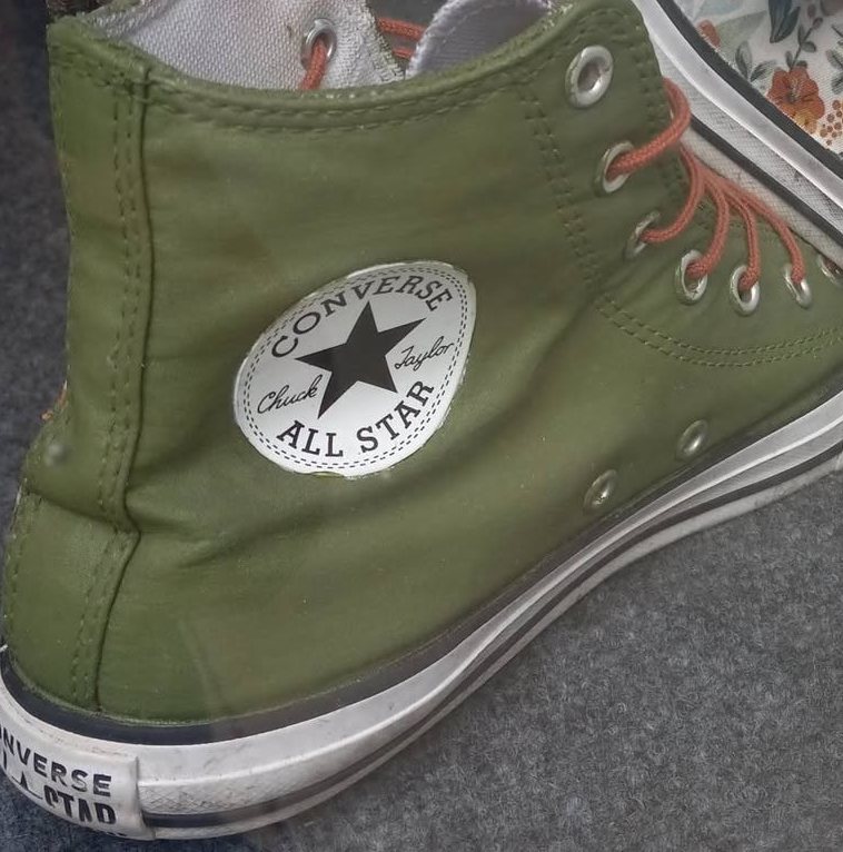 A worn olive-green canvas high-top sneaker with orange laces and a white rubber toe cap, on grey felt.