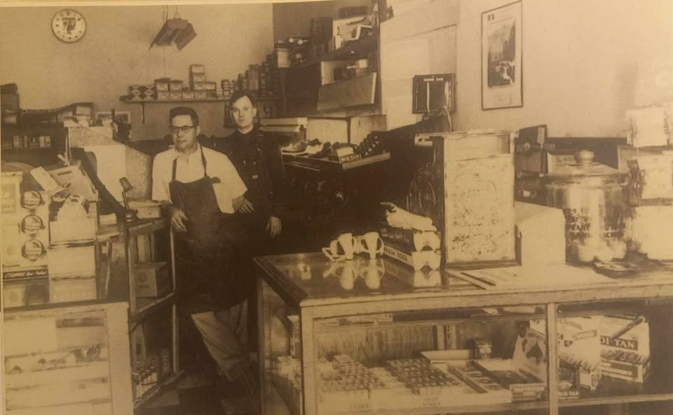 A sepia photograph from the middle of the last century: two men in a shoe repair shop, one in a leather apron, standing between a glass display case of polishes and laces and a wall of shoe boxes.